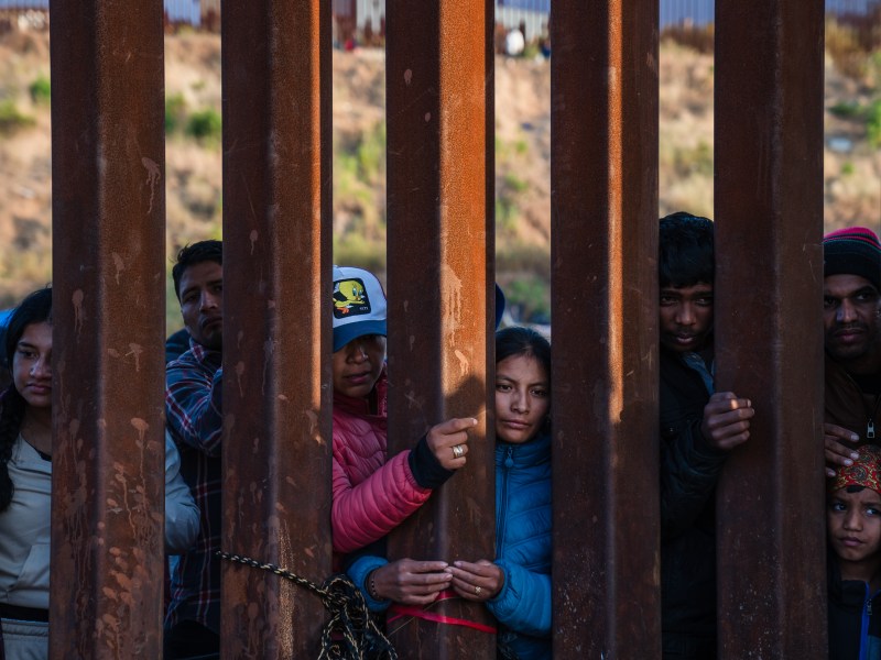 Asylum seekers can be seen through the border wall waiting for snacks and fruit that were given out by volunteers in San Ysidro on May 11, 2023.