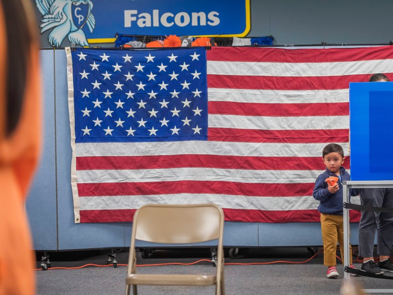 A little boy stands near a voting both, while his guardian votes on Tuesday, Nov. 5, 2024. / Photo by Vito di Stefano for Voice of San Diego